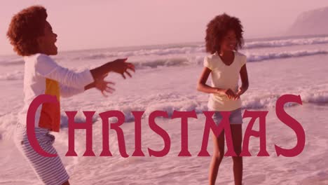 Animation-Eines-Weihnachtstextes-über-Einer-Glücklichen-Afroamerikanischen-Mutter-Und-Kindern,-Die-Am-Sonnigen-Strand-Spielen