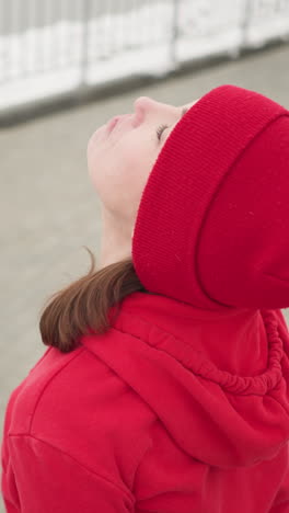 mujer con capucha roja levantando la cabeza hacia atrás y hacia adelante lentamente en una escena al aire libre nevada con barandilla de hierro en el fondo, evocando movimiento pacífico y serenidad en la fría atmósfera de invierno
