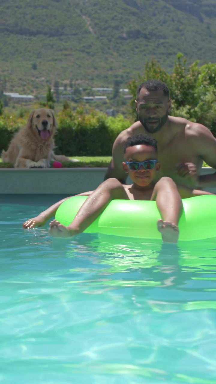 Vertical video: Playing shirtless dad steadying son in home pool after climbing neon tube dog lying