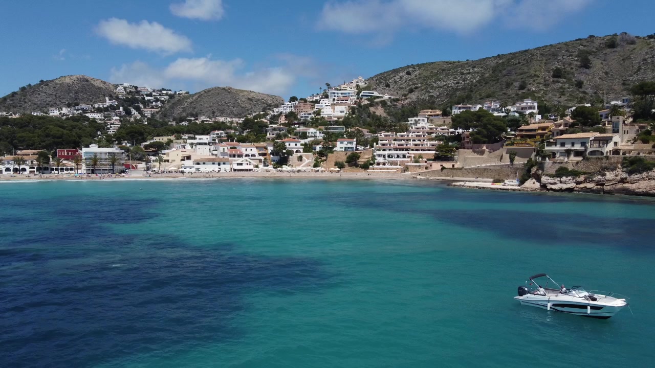 Ripresa cinematografica con drone che si allontana dalla spiaggia di El Portet a Moraira, Spagna, rivelando ville di lusso incastonate sulle colline e yacht bianchi ancorati nella baia turchese cristallina