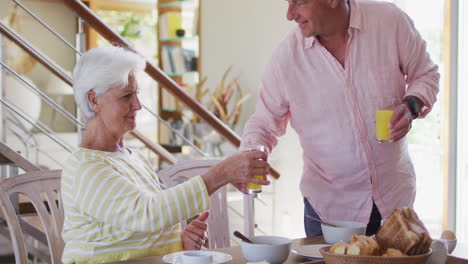 Pareja-Caucásica-Mayor-Tostando-Vasos-De-Jugo-Mientras-Desayunan-Juntos-En-Casa