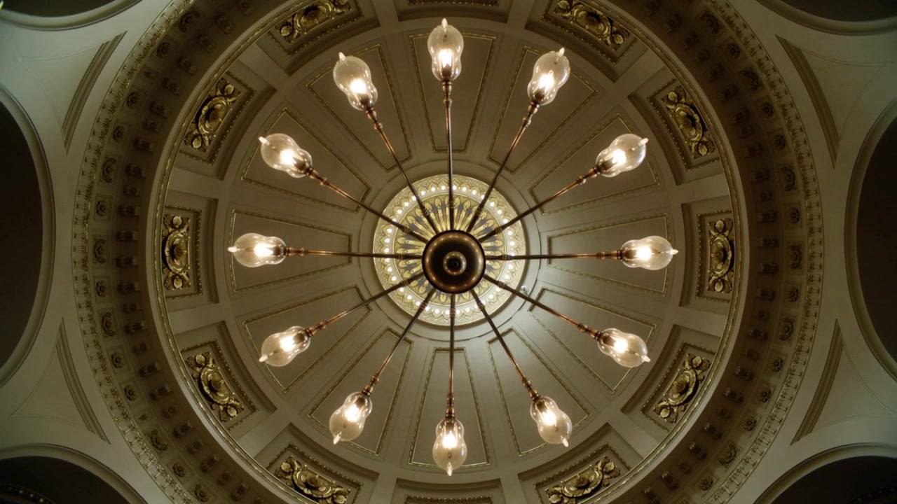 La telecamera inizia un avvicinamento verso il lampadario centrale in una grande sala, rivelando un medaglione dorato.
