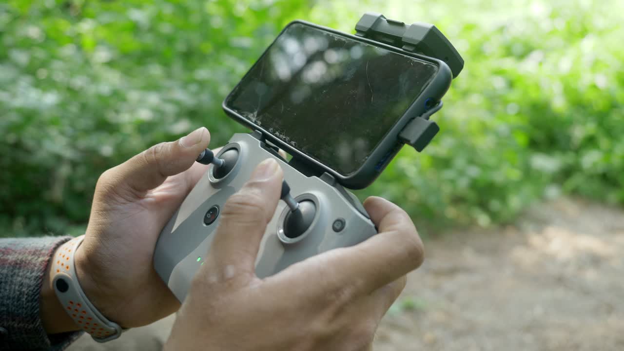 Closeup of hand of drone pilot using DJI drone remote controller with a smartphone attached in a forest landscape for wildlife surveillance, India