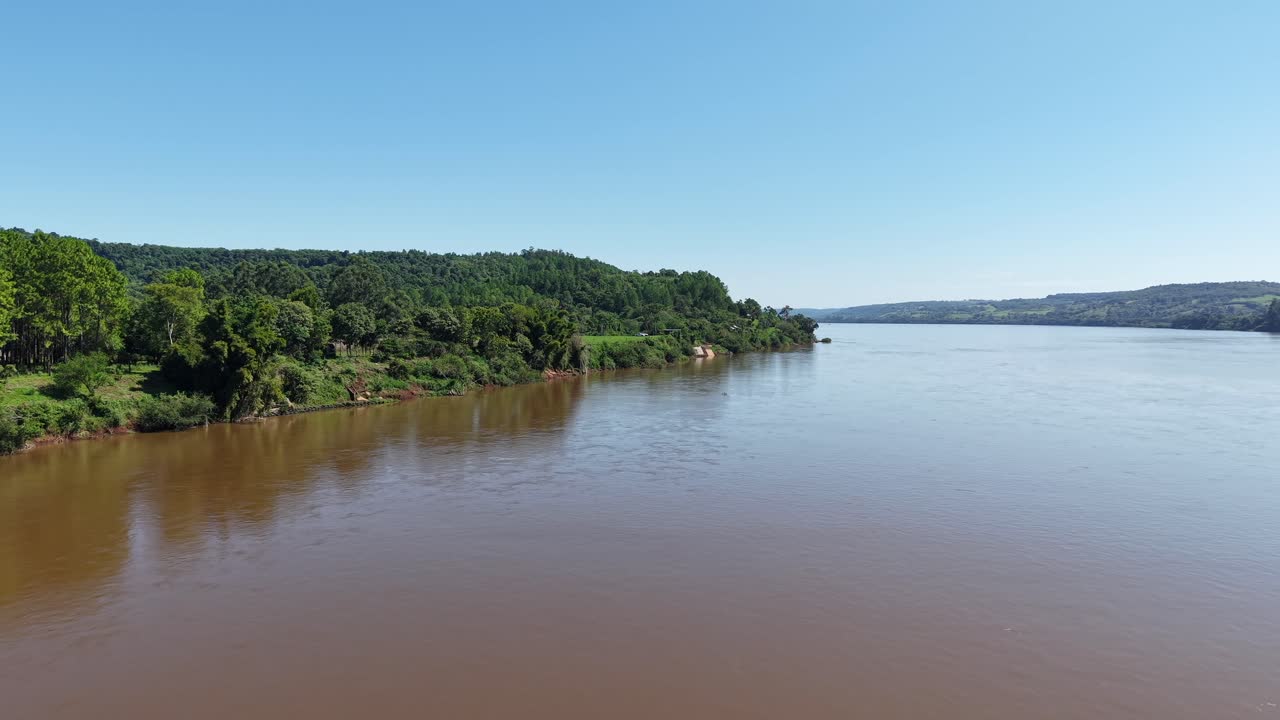 Aerial drone view flying over the wide muddy Rio Uruguay near Panambi Misiones Argentina showing lush green forest banks under a clear blue sky
