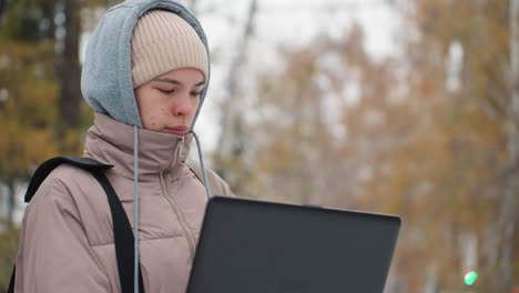 young woman bundled in hooded winter coat and beanie intently focused on laptop she is holding while standing outside on snowy ground with soft autumn colors in background