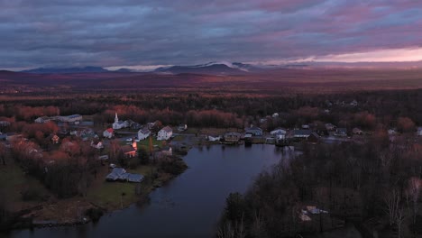 Luftaufnahmen,-Die-Bei-Sonnenaufgang-Im-Spätherbst-Rückwärts-Von-Einer-Kleinen-Gemeinde-Am-Ufer-Des-Sees-Wegfliegen,-Mit-Nebligen-Bergen-Im-Hintergrund