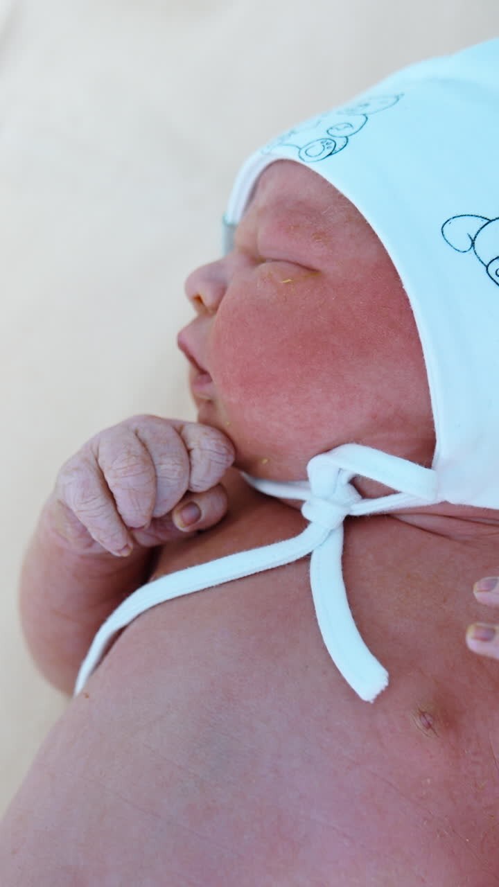 Caucasian newborn naked in only white cap lies on the table. Tiny child yawns and cries. Close up. Vertical video