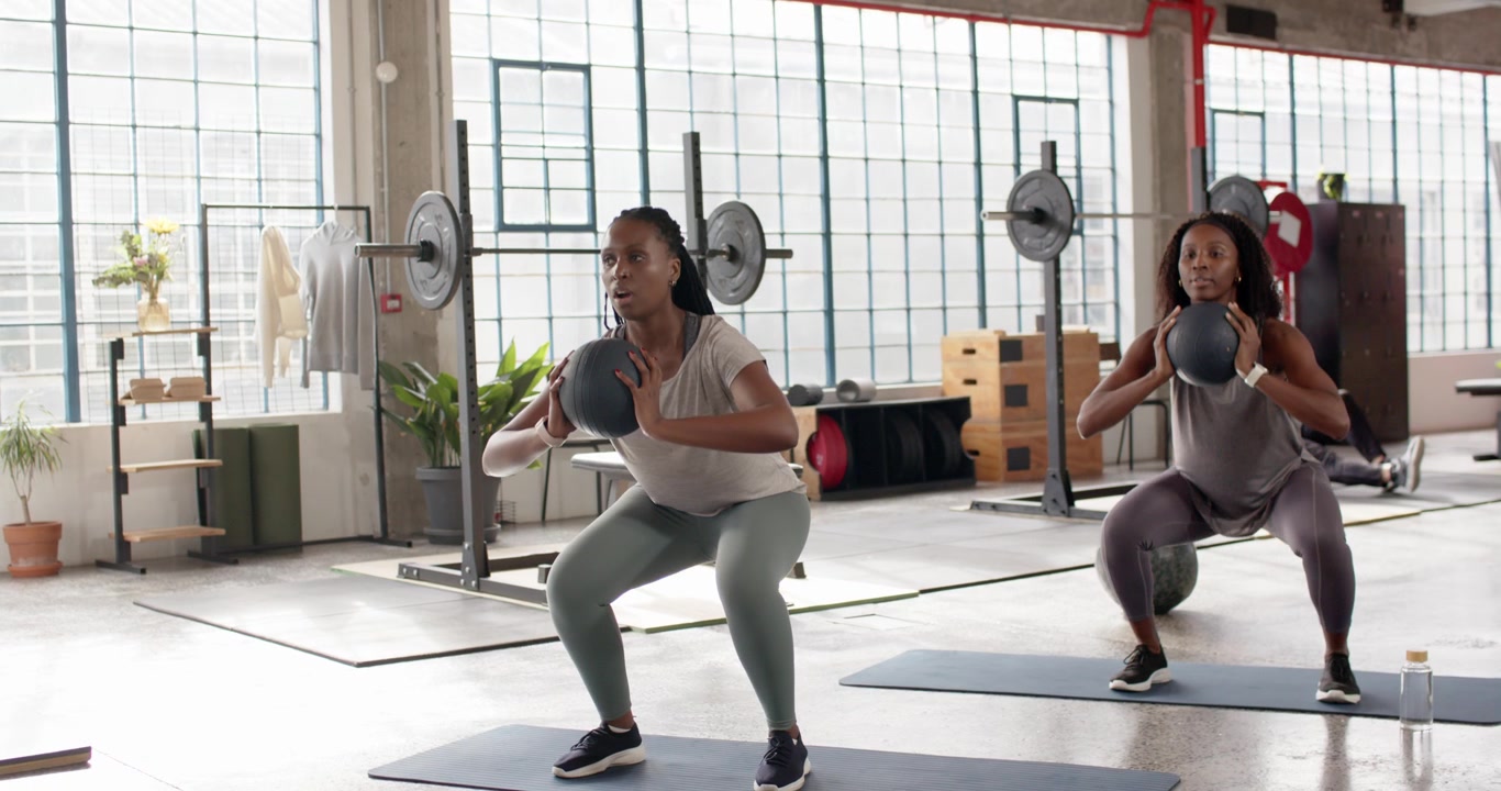 Donne afroamericane che eseguono squat frontali con palla medica in una palestra loft, quella a sinistra guida l'allenamento.