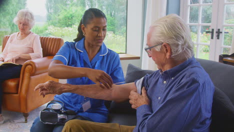 senior man at home in lounge having blood pressure taken by female care worker in uniform