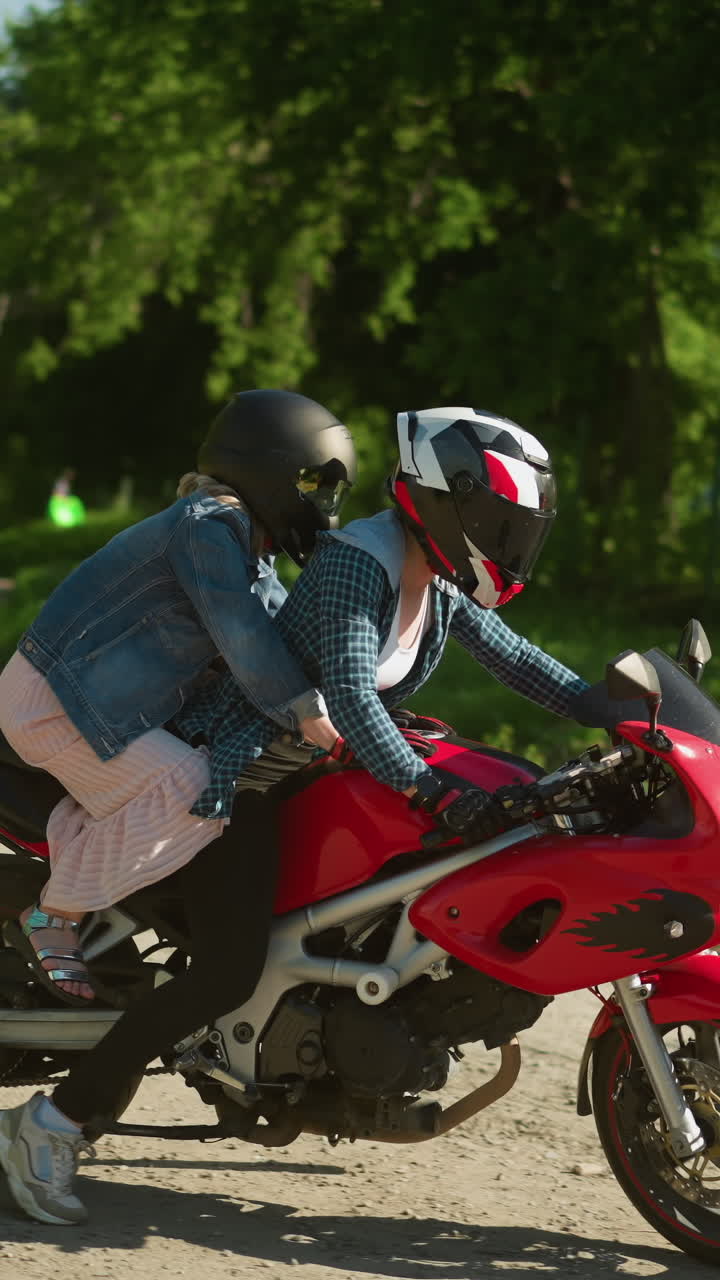 dos hermanas montan una bicicleta eléctrica roja, ambas con cascos, mientras conducen en un camino pavimentado rodeado de árboles, se puede ver una vista borrosa de los edificios en el fondo