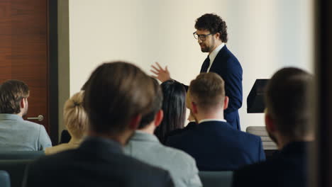 caucasian businessman wearing glasses and formal clothes speaking at a conference in front of many people