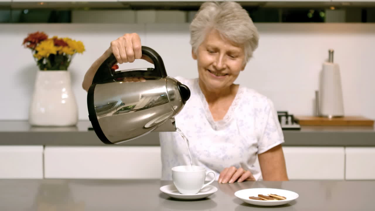 mujer jubilada vertiendo agua hirviendo de la tetera en la taza en la cocina
