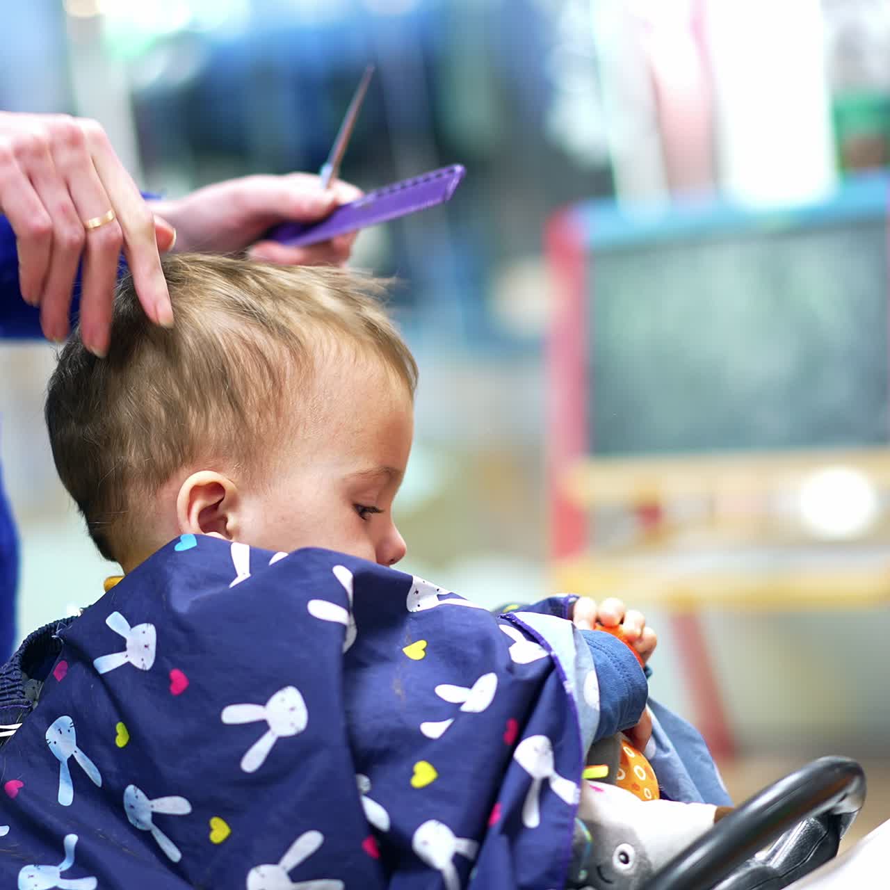 Ein bezaubernder kleiner Junge ist mit einem Spielzeug beschäftigt, während der Friseur seine Haare schneidet. Ein bezauberndes einjähriges Kind lässt sich im Salon die Haare schneiden
