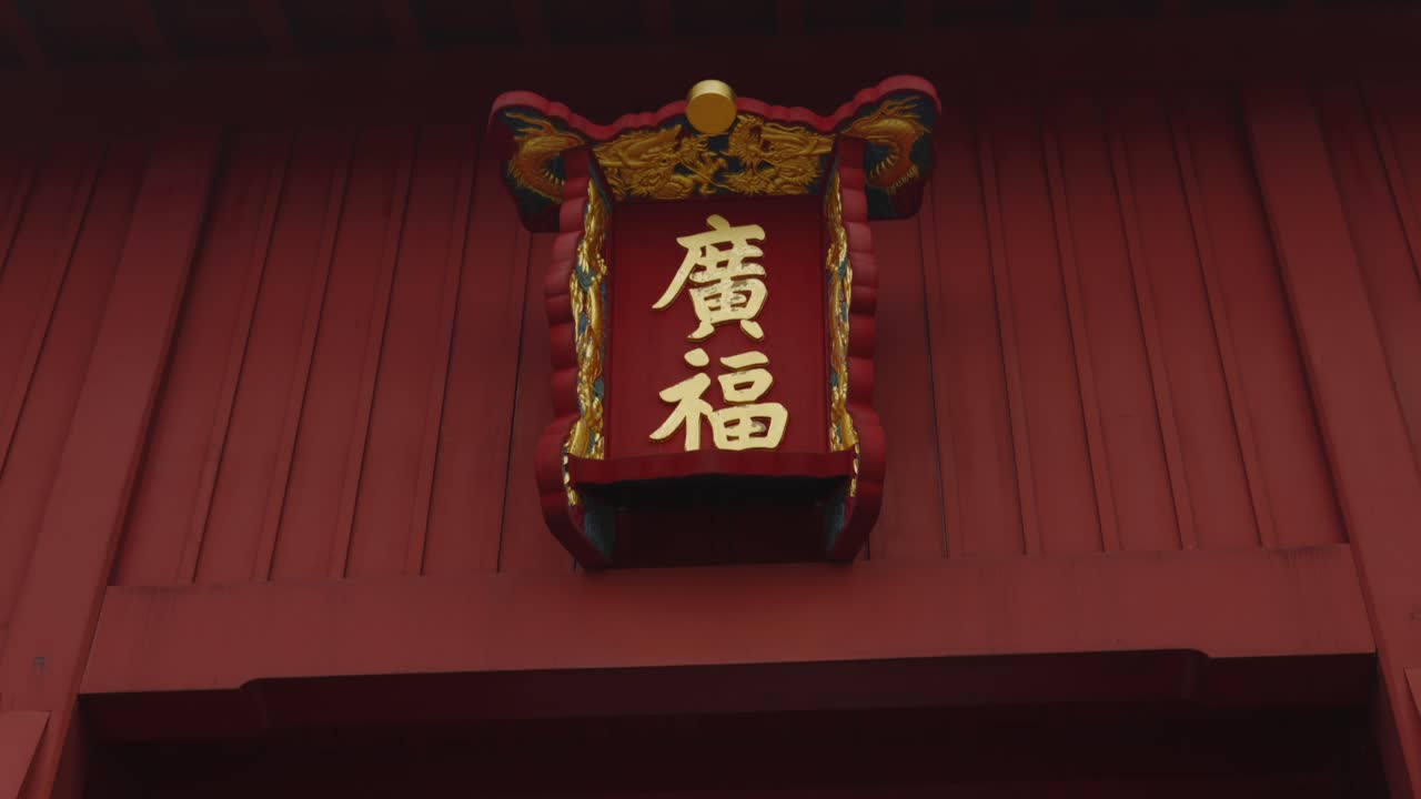 Et dynamisk tilt-skud bevæger sig ned langs den detaljerede røde struktur af Shuri Slot og afslører det gyldne Kanji-skilt på hovedporten. Den traditionelle okinawiske arkitektur er slående mod den overskyede himmel.