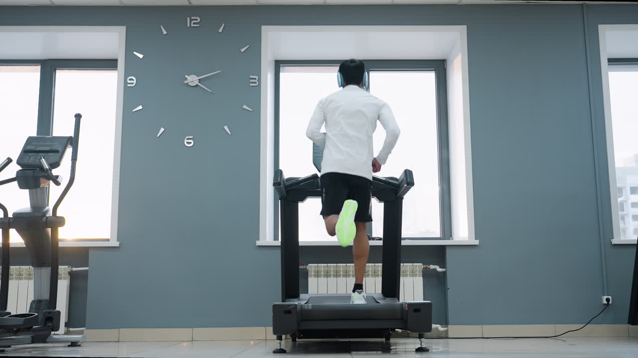 rear view of runner adjusting white top on treadmill in modern gym with elliptical trainer and strength machine on each side sunlight glow through window casting warm highlights on blue walls