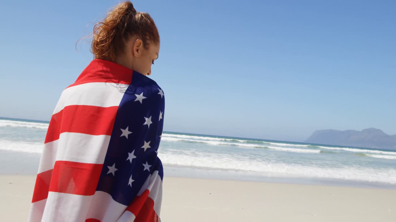 mujer envuelta en bandera estadounidense. de pie en la playa 4k