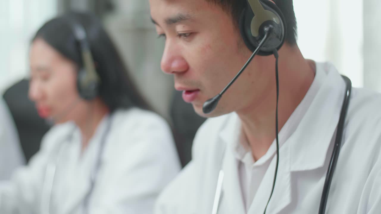 Close Up Of An Asian Man Doctor Wearing Headsets Working As Call Center Agent Speaking To Customer On The Call And Typing During Woking With Colleagues At The Office
