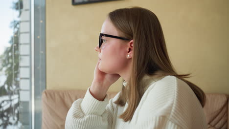 lonely woman seated indoors stirs coffee slowly while resting hand under jaw, lost in thought, soft daylight from window and wall art add warmth to quiet and reflective cafe moment