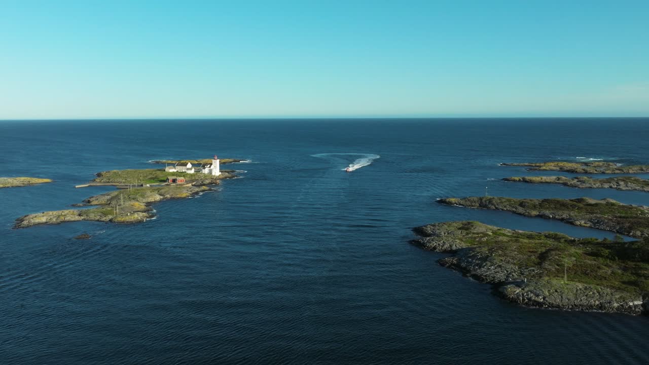 A lighthouse stands on a small island surrounded by calm waters. A boat moves along the surface, creating ripples against the clear blue sky, capturing a peaceful moment in nature