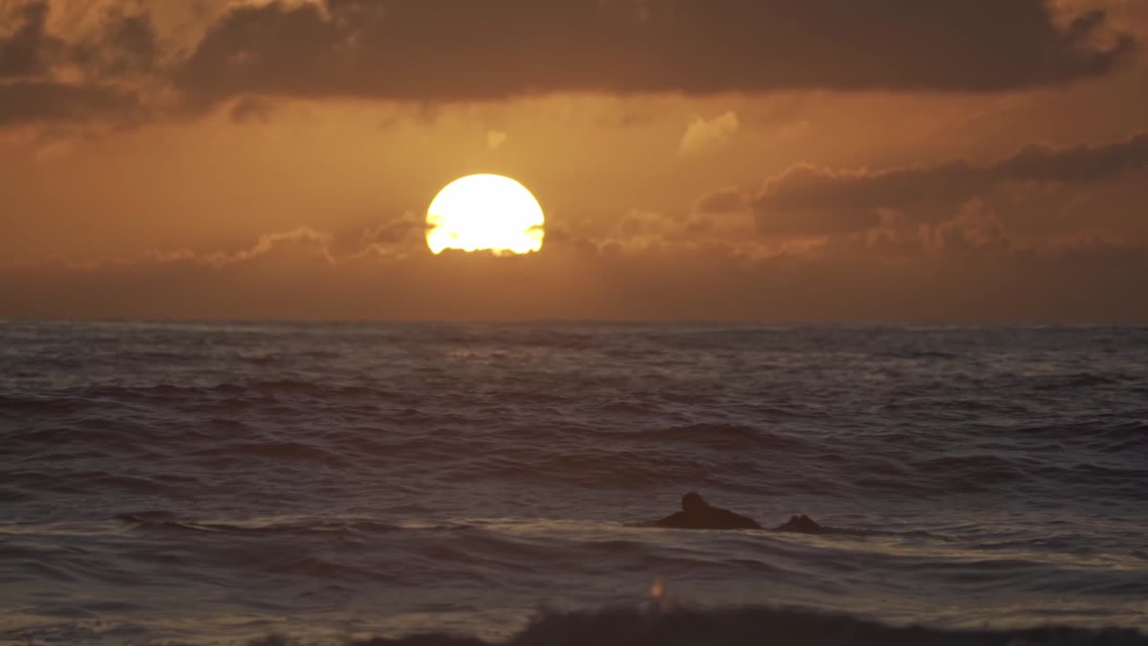 A captivating wide shot of surfers in the Pacific Ocean during a vibrant sunset in Oaxaca, Mexico. The large, radiant sun dips towards the horizon, casting a golden glow across the sky