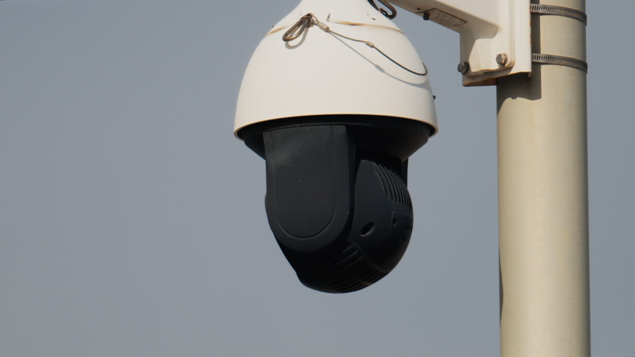 Close up of a round, black and white surveillance camera mounted on a white pole with the cloudy sky on the background