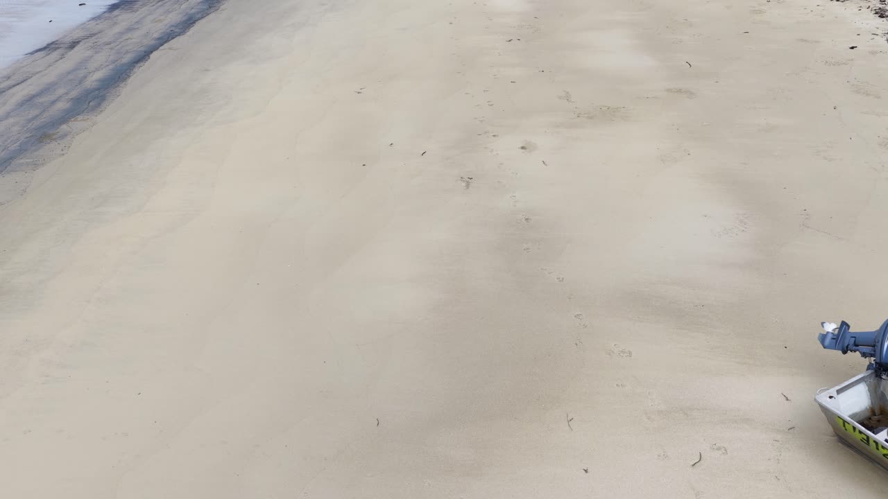 Drone glides toward aluminum motorboat on empty tropical beach, soft daylight, wide perspective