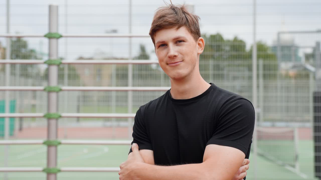 Young Caucasian Man In Black T-Shirt With His Arms Crossed, Smiling At The Camera On An Outdoor Sports Court. - medium shot