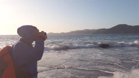 young adult man taking pictures of sunlit seascape on baker beach san francisco