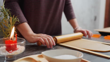 baker hands rolling brown dough with wooden rolling pin, left wrist adorned with pink beaded bracelet, slight blur in background featuring candle, kitchen tools