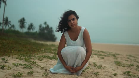 Woman-in-white-dress-sitting-thoughtfully-on-a-tropical-beach-at-dusk,-close-up