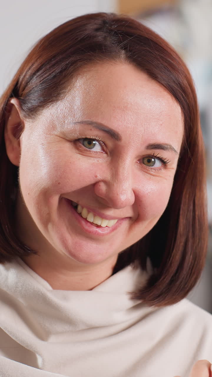 Mujer caucásica recortando una cinta con unas tijeras, manos concentradas usando unas grandes cizallas negras, suéter beige, taller de fondo, sonrisa suave, corte de pelo bob, primer plano de las manos y la cara, momento creativo satisfactorio.