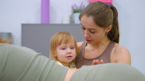 a touching scene of a mother and daughter holding each other warmly in a gym, with the toddler playfully interacting with a woman working out in the background