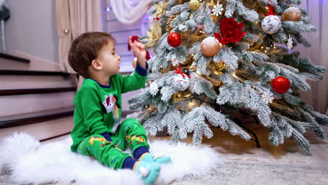cute dark-haired baby boy in green suit sits near the christmas tree. lovely kid touching the toys on the tree.