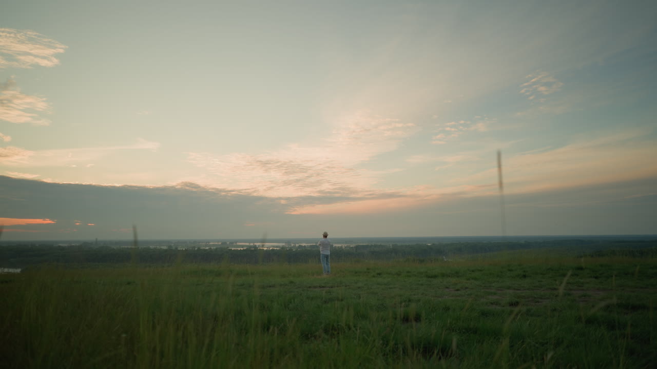 색 셔츠, 모자, 청바지를 입은 남자가 멀리서 보이는데, 해가 지는 동안 조용한 호수 의 풀에서 상하고 서 있다. 넓은 하늘과 조용한 배경은 평화의 느낌을 불러일으킨다.