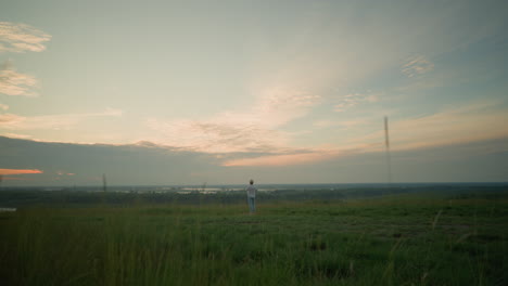 a distant view of a man dressed in a white shirt, hat, and jeans, standing contemplatively in a grassy field by a tranquil lake at sunset. the expansive sky and serene setting evoke a sense of peace