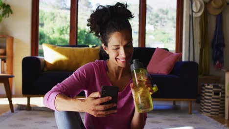 Mixed-race-woman-drinking-water-and-using-smartphone-while-sitting-on-yoga-mat-at-home