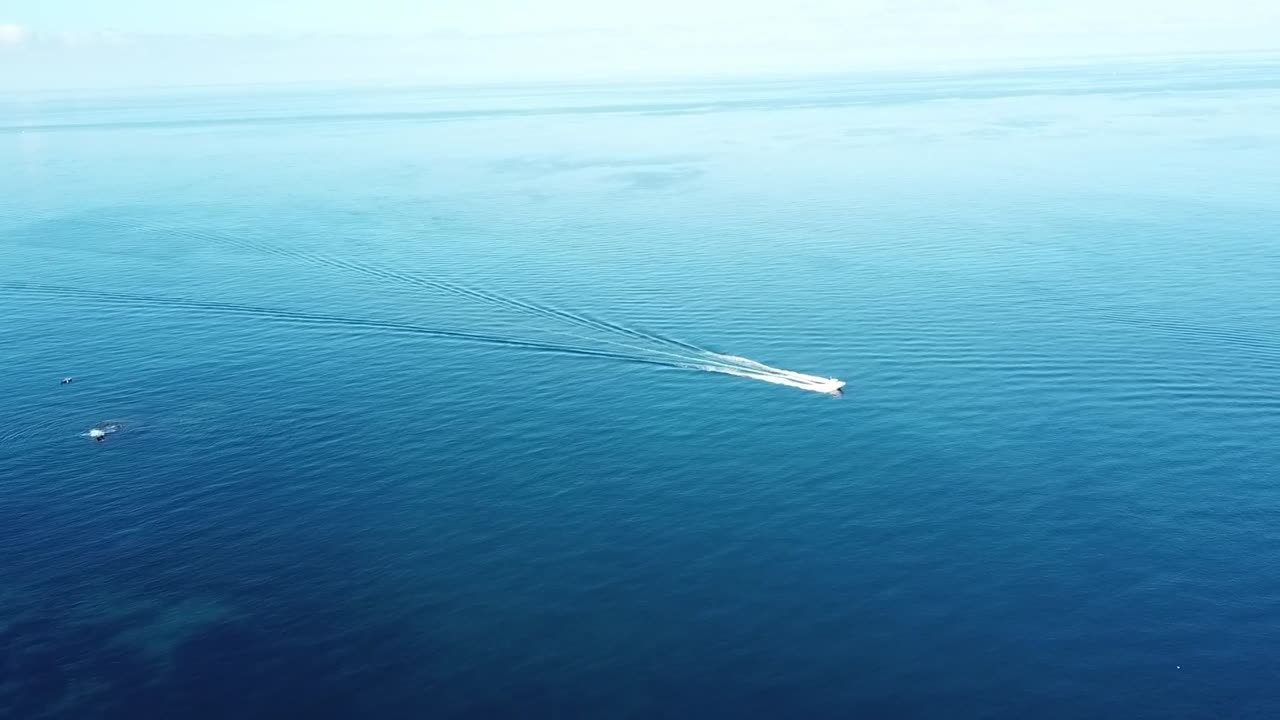 Cinematic drone shot tracking a small white boat leaving a long white wake trail as it speeds through deep blue waters
