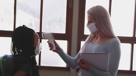 female teacher wearing a face mask measuring temperature of a girl in class at school