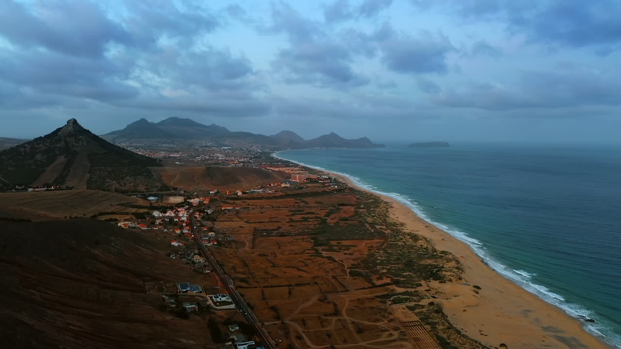 Plain shoreline with a town located on it. Dramatic cloudscape in the sky above the scenery. Mountains silhouettes at backdrop. Madeira Island coast at the Atlantic Ocean.