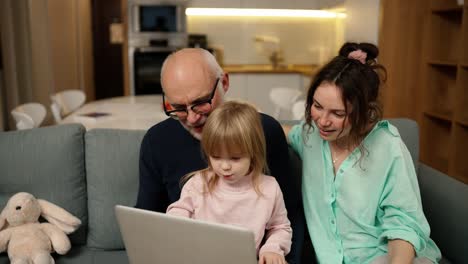 Granddaughter,-daughter-teaching-senior-grandparent-using-laptop-sitting-on-sofa-in-living-room