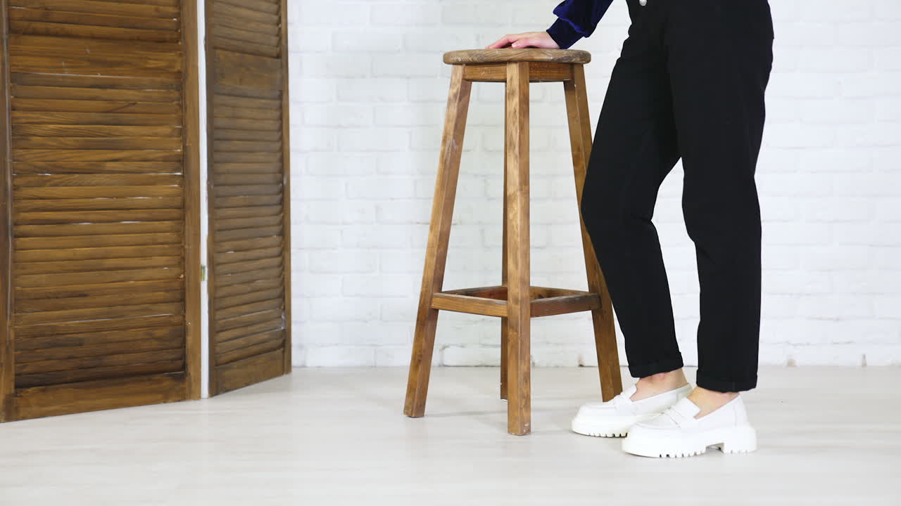 Woman in black jeans comes to a wooden stool showing white shoes. Footwear presentation at white backdrop.