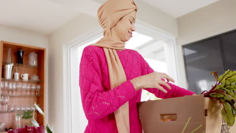 happy biracial woman in hijab with grocery shopping in kitchen at home with copy space, slow motion