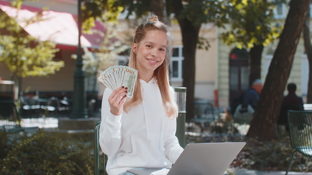Happy rich woman using laptop winner victory holding cash money in dollar banknotes sitting on chair