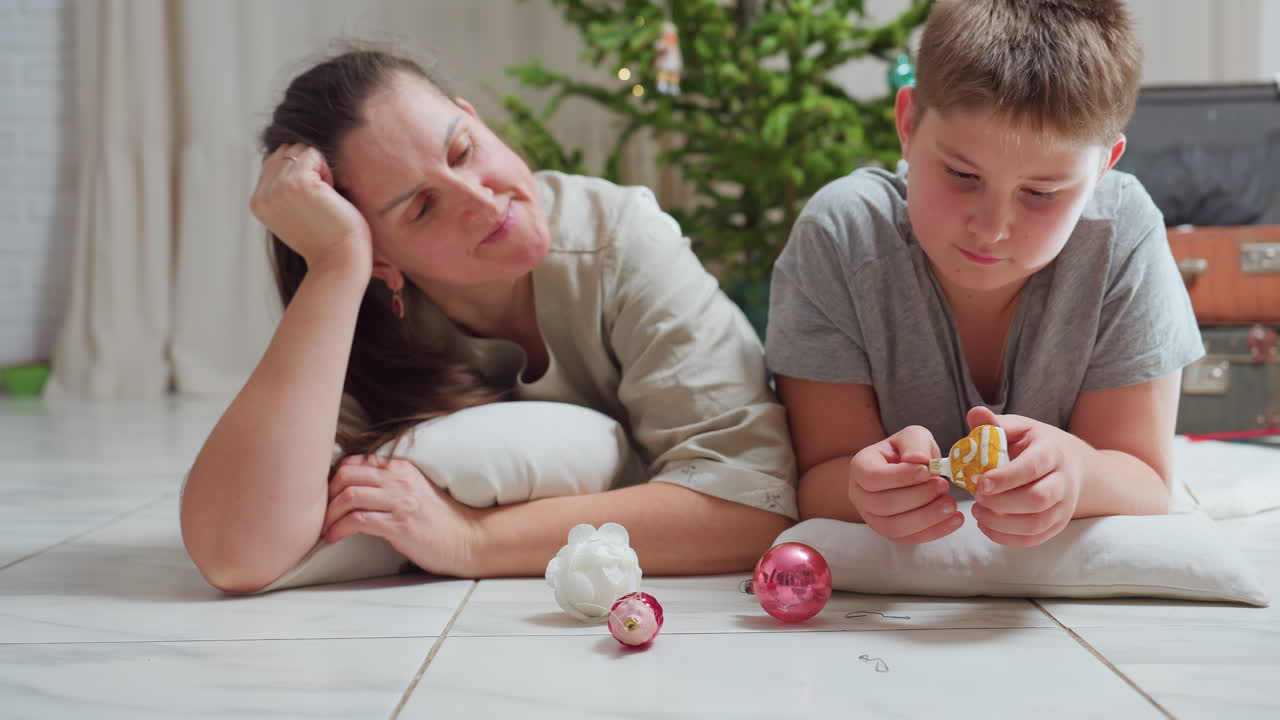 mom and son lying on marble floor close together surrounded by colorful christmas ornaments smiling warmly cozy festive home atmosphere decorated christmas tree in background bright joyful holiday