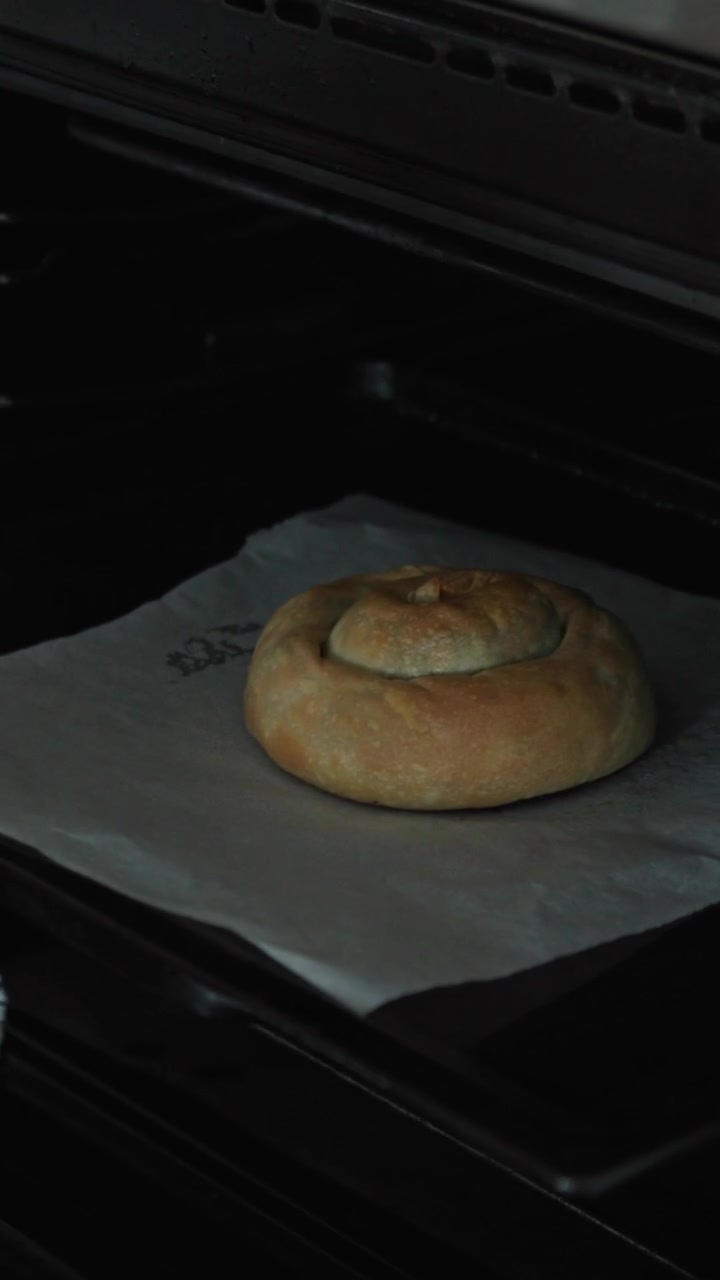 Primer plano de una spanakopita en espiral recién horneada siendo retirada del horno sobre papel de horno usando un paño de cocina a rayas, ambiente cálido de cocina casera, toma vertical.