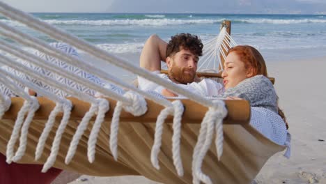 Front-view-of-young-romantic-caucasian-couple-relaxing-in-hammock-at-beach-on-a-sunny-day-4k