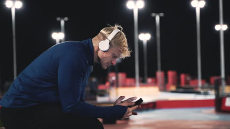 lächelnder sportler sitzt im park und hört musik mit bluetooth-kopfhörern, schreibt sms auf sein handy und schaut sich um, während er nachts während seiner trainingseinheit eine pause macht 2