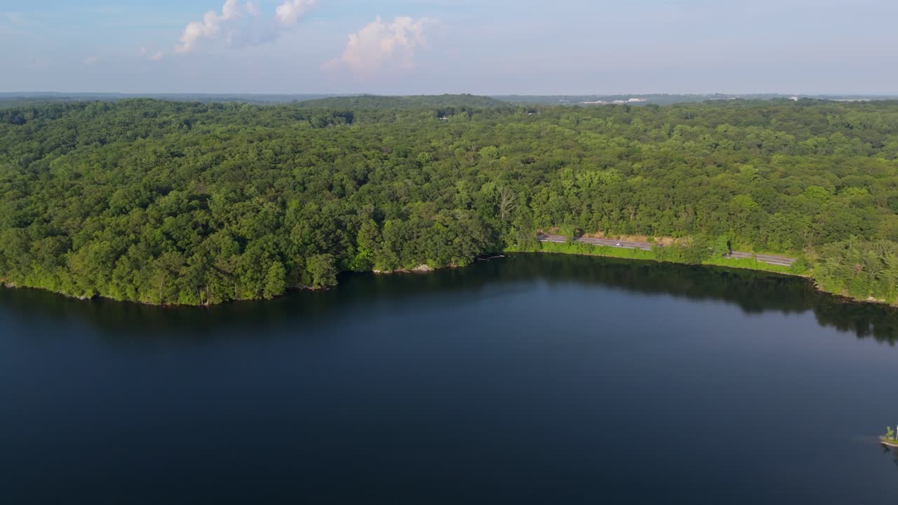 Calm and deep blue water of Kensico Reservoir, vast green forest and scenic road surround lake, Westchester County, New York, USA. Aerial drone lateral view