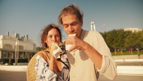 Porträt-Eines-Glücklichen-Paares,-Mann-Und-Frau,-Die-Morgens-Hotdogs-An-Einem-Modernen-Strand-Essen.-Blonder-Mann-In-Leichter-Kleidung-III.-Braunhaariges-Mädchen-Isst-Morgens-Hotdogs-An-Einem-Sonnigen-Strand