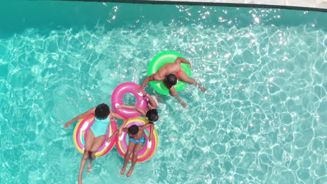 Aerial-of-african-american-parents,-son-and-daughter-sitting-on-inflatable-in-sunny-pool,-copy-space